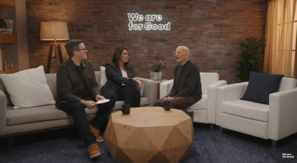 Three people sit talking in a cozy ASL Studios setting with a brick wall, plants, and neutral armchairs. A neon sign above them reads 